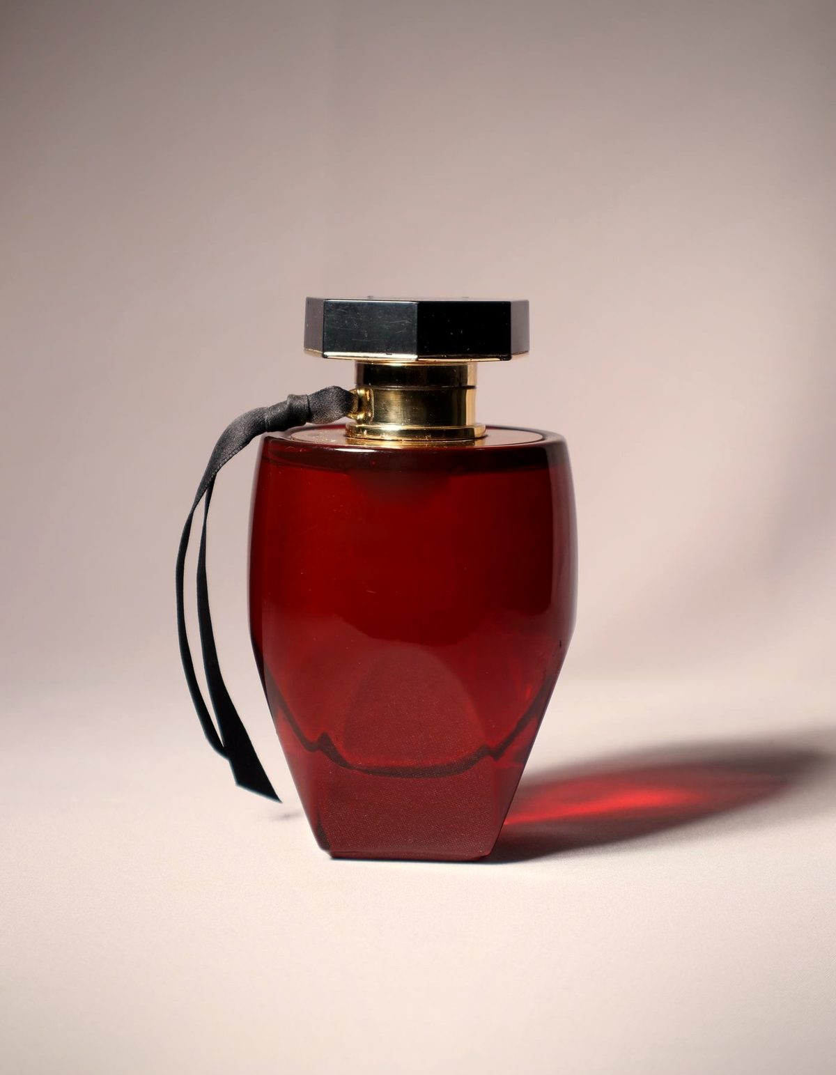 Translucent deep-red glass perfume bottle with a gold collar and faceted black cap, lit softly against a warm neutral background
