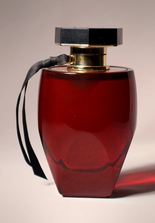 Translucent deep-red glass perfume bottle with a gold collar and faceted black cap on a warm neutral background
